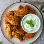 A plate of seasoned chicken wings garnished with herbs, served with a bowl of creamy white dipping sauce topped with mint, on a wooden surface.