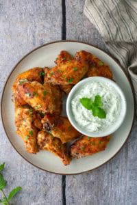 A plate of seasoned chicken wings garnished with herbs, served with a bowl of creamy white dipping sauce topped with mint, on a wooden surface.