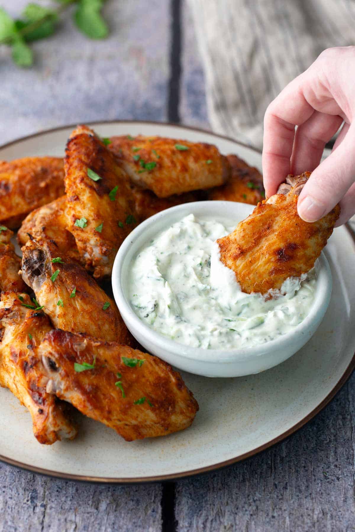 A hand dipping a seasoned chicken wing into a bowl of creamy white dipping sauce, with more chicken wings arranged on a plate.