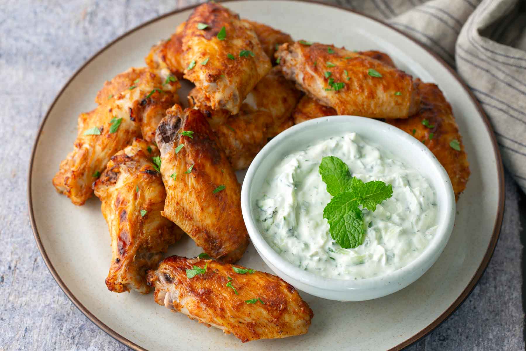 A plate of seasoned chicken wings garnished with herbs, served with a bowl of creamy herb dip topped with a mint sprig.