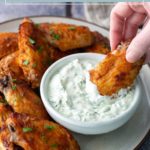 A hand dips a baked chicken wing into a bowl of creamy white dip, with several more wings arranged on a plate.
