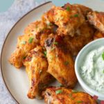A plate of baked chicken wings garnished with herbs, served with a bowl of creamy dipping sauce on the side.