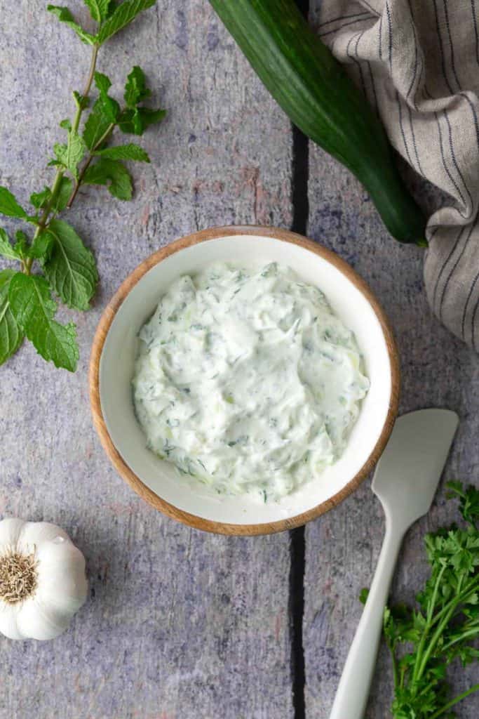A bowl of creamy tzatziki dip sits on a gray surface next to a cucumber, mint, garlic, parsley, and a white spatula.