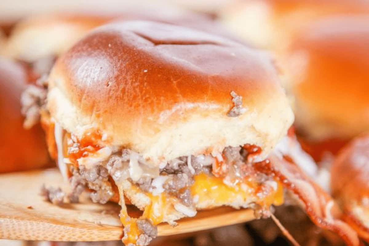 Close-up of a cheeseburger slider with ground beef, melted cheese, bacon, and sauce on a soft bun.