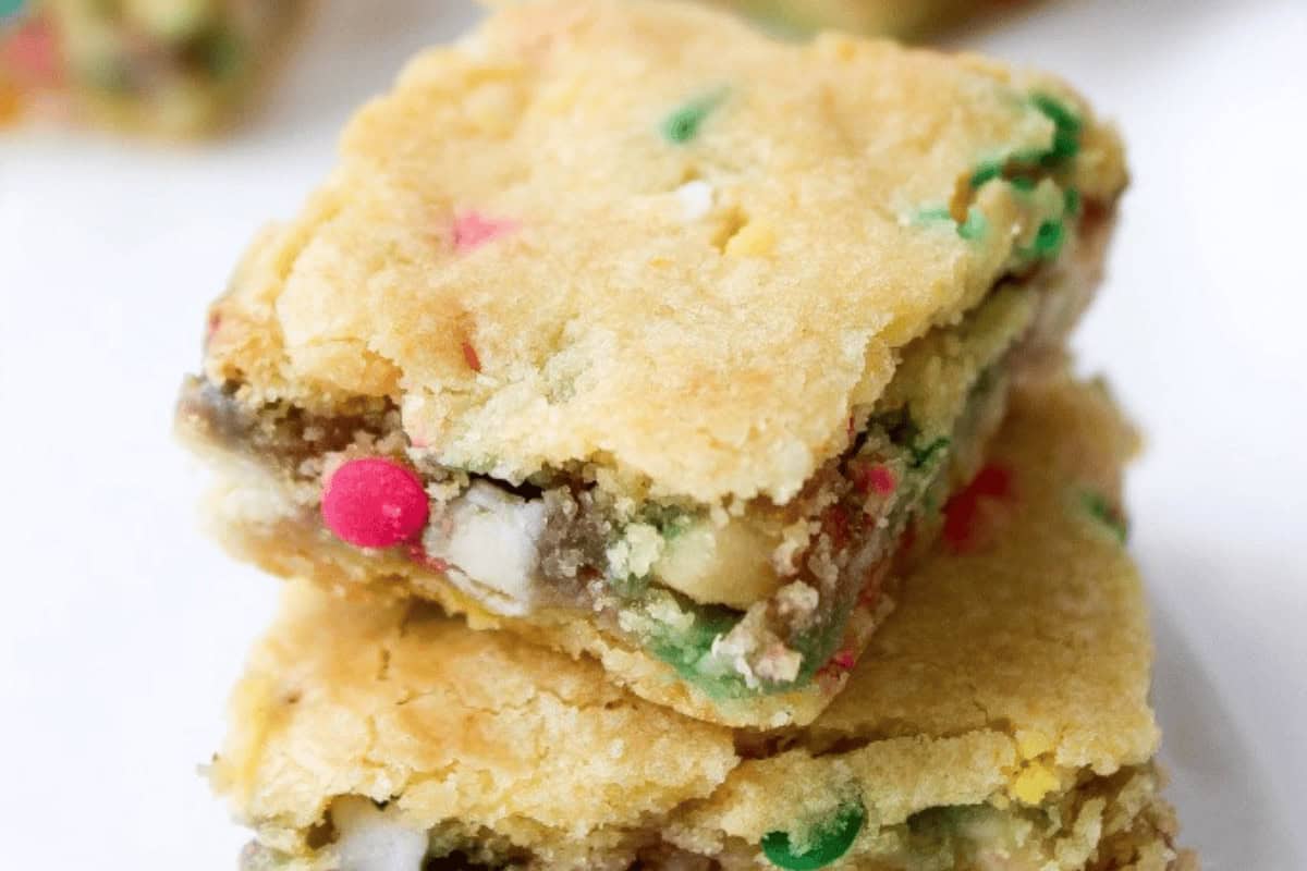 Two stacked cookie bars with a golden crust, containing colorful candy pieces and white chocolate chips, on a white background.