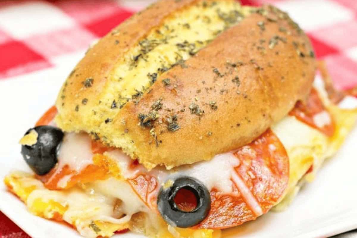 A sandwich on an herb-topped bun filled with melted cheese, pepperoni slices, and black olives, served on a white plate.