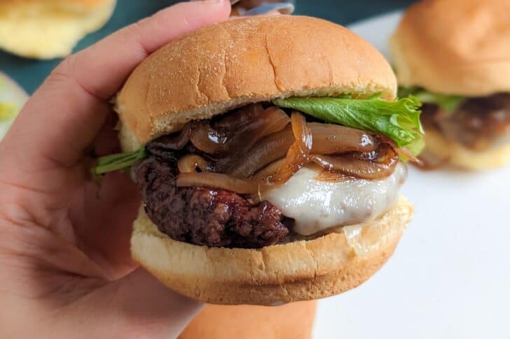 A hand holding a hamburger with lettuce, melted cheese, caramelized onions, and a beef patty in a bun.