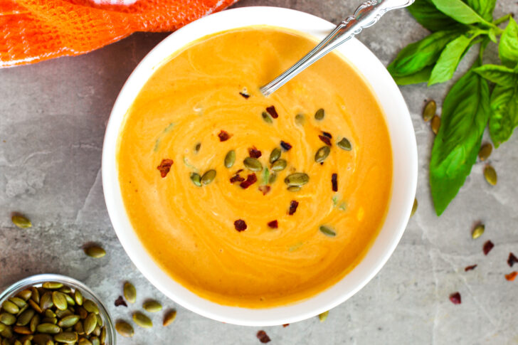 A bowl of creamy orange soup garnished with pumpkin seeds and chili flakes, with a spoon inside. Fresh basil leaves and a small dish of pumpkin seeds are nearby on the table.