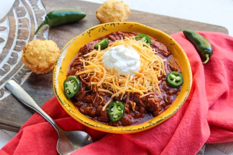 A bowl of chili topped with shredded cheese, sliced jalapeños, and sour cream, served with two cornbread muffins and a spoon on a wooden board with a red napkin.