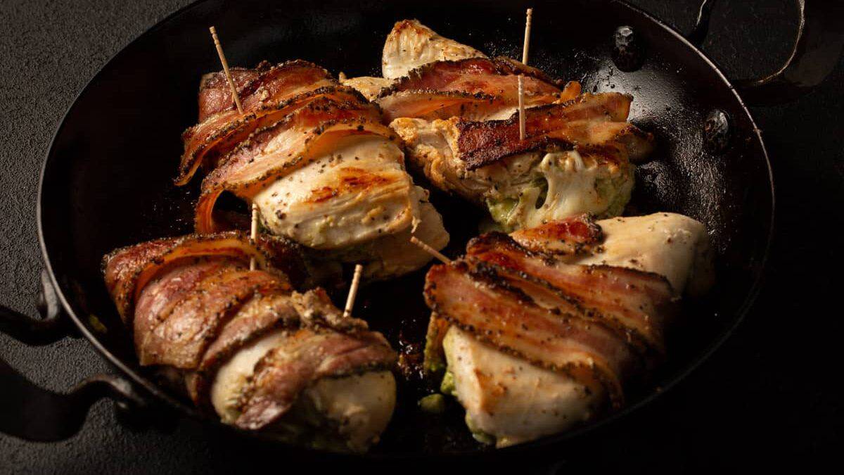 Chicken breasts stuffed with vegetables and wrapped in bacon, secured with toothpicks, are cooking in a black pan.