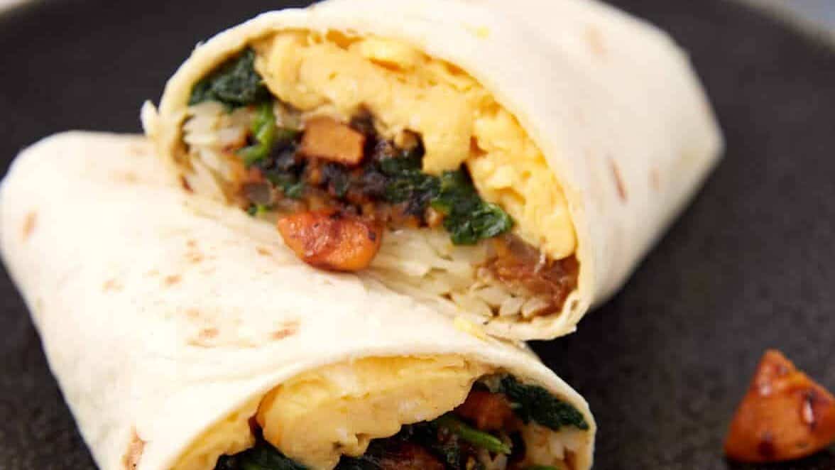 A breakfast burrito cut in half, showing scrambled eggs, sausage, rice, spinach, and other fillings wrapped in a flour tortilla.