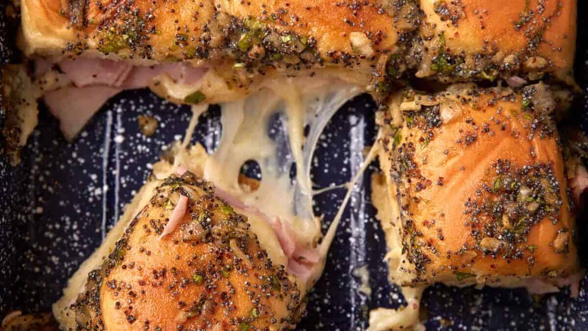 Close-up of baked ham and cheese sliders with poppy seed topping in a baking dish, showing melted cheese stretching between sandwiches.