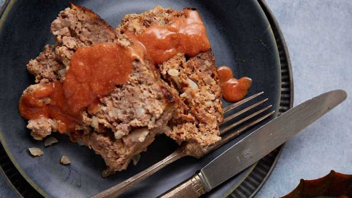 Two slices of meatloaf with tomato sauce on a dark plate, served with a fork and knife.