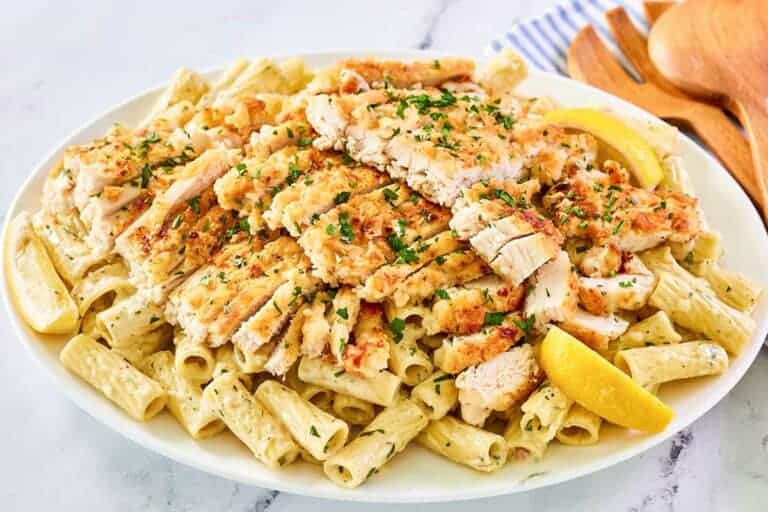 A plate of rigatoni pasta in creamy sauce topped with sliced grilled chicken breast, garnished with chopped herbs and lemon wedges.