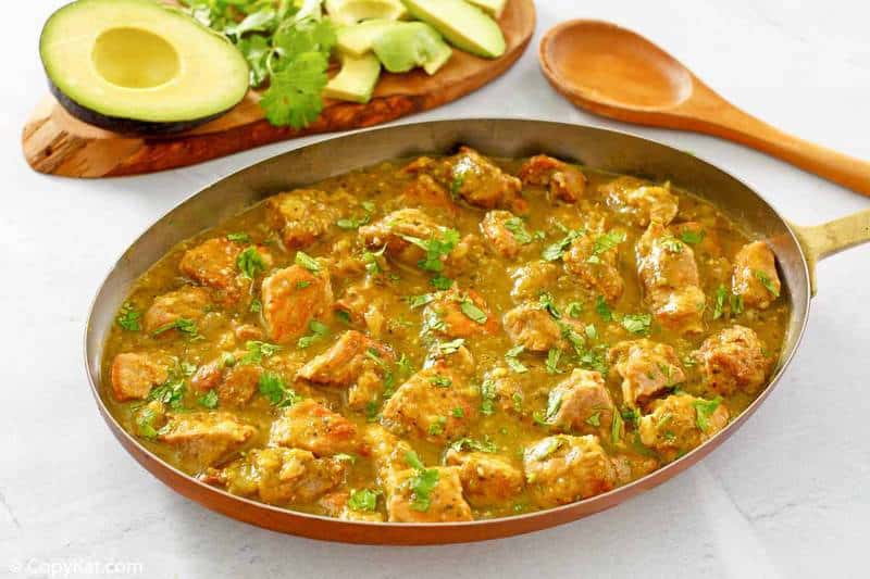 A skillet filled with pork in green chili sauce, garnished with cilantro, sits next to a wooden spoon and a cutting board with sliced avocado and cilantro.