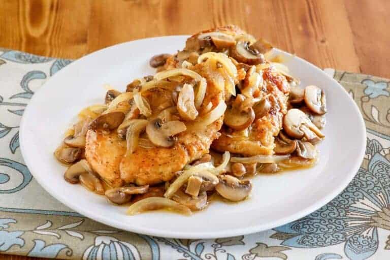 A white plate with cooked chicken breasts topped with sautéed mushrooms and onions, set on a patterned cloth on a wooden table.