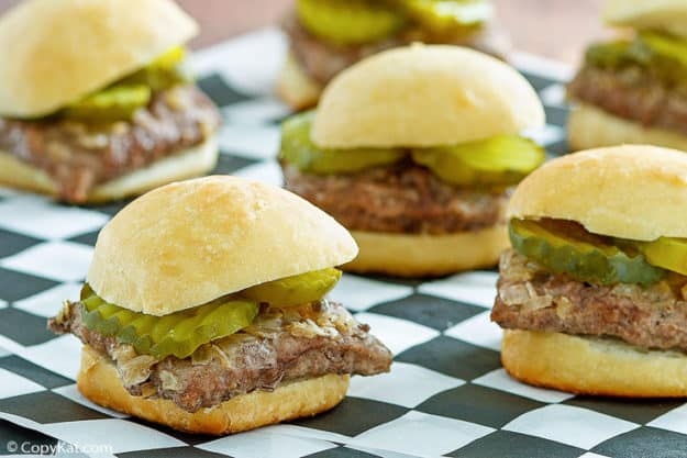 Mini beef sliders topped with pickles and chopped onions are arranged on a black and white checkered surface.