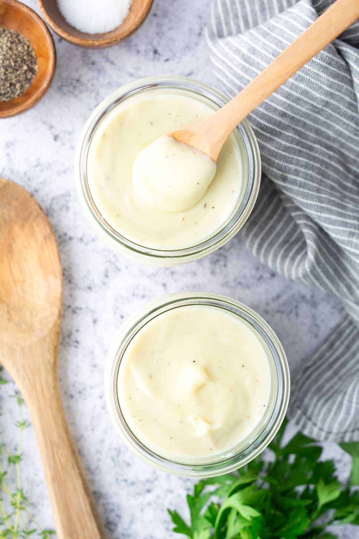 Two glass jars filled with creamy white sauce sit on a marble surface, surrounded by a wooden spoon, herbs, and a striped cloth; a small wooden spoon rests in one jar.