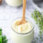 A small glass jar filled with creamy white sauce, with a wooden spoon inserted, sits on a light surface next to fresh herbs.