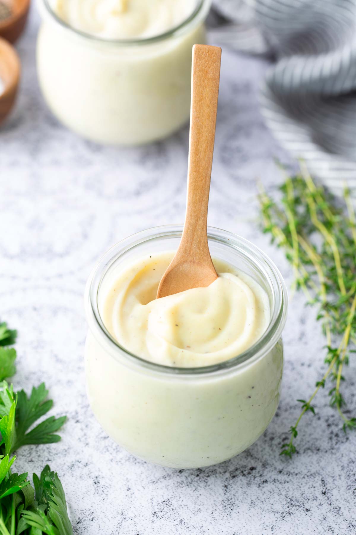 A small glass jar filled with creamy white sauce, with a wooden spoon inserted, sits on a light surface next to fresh herbs.
