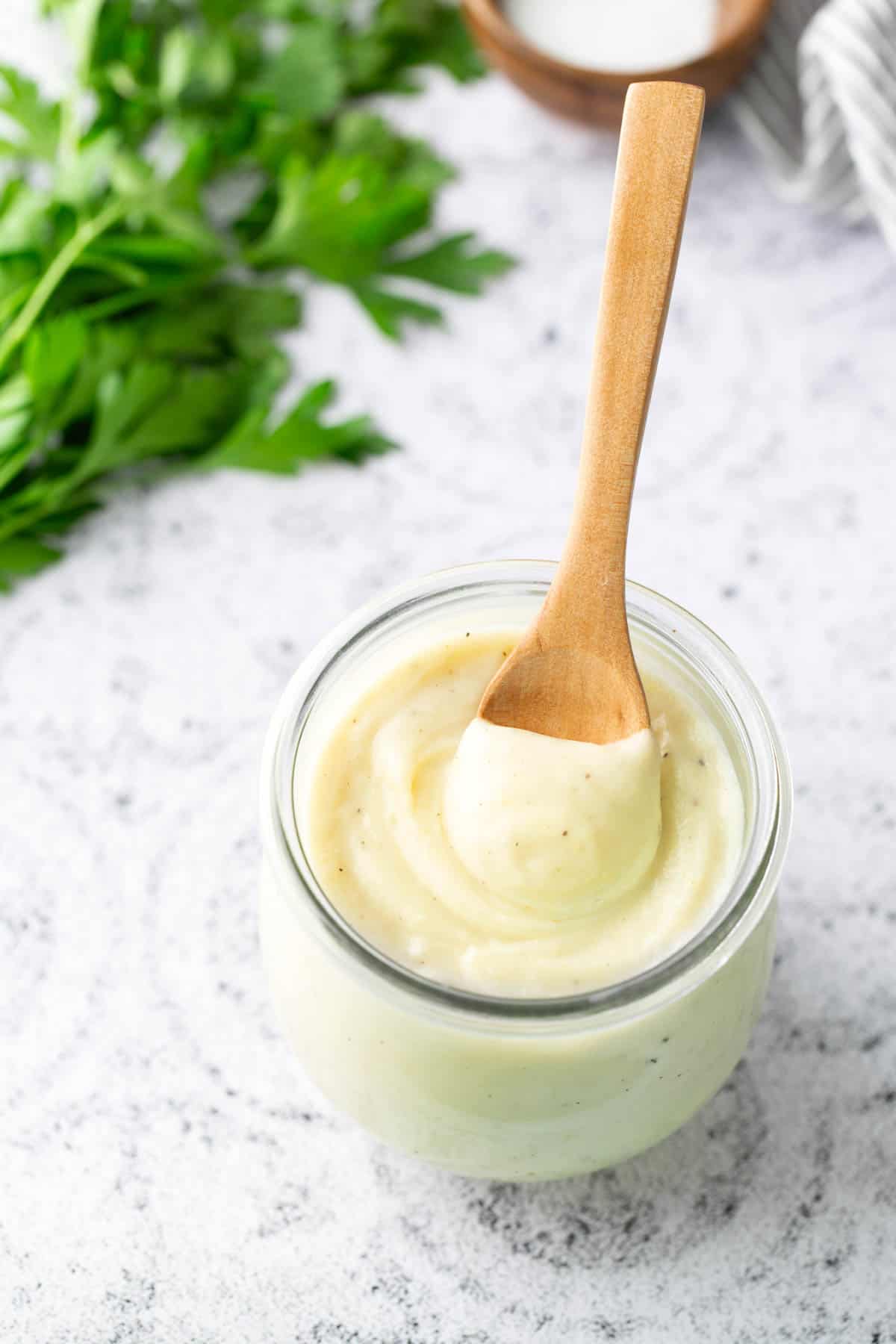 A glass jar filled with creamy mashed potatoes, topped with a wooden spoon. Fresh parsley is in the background on the left.