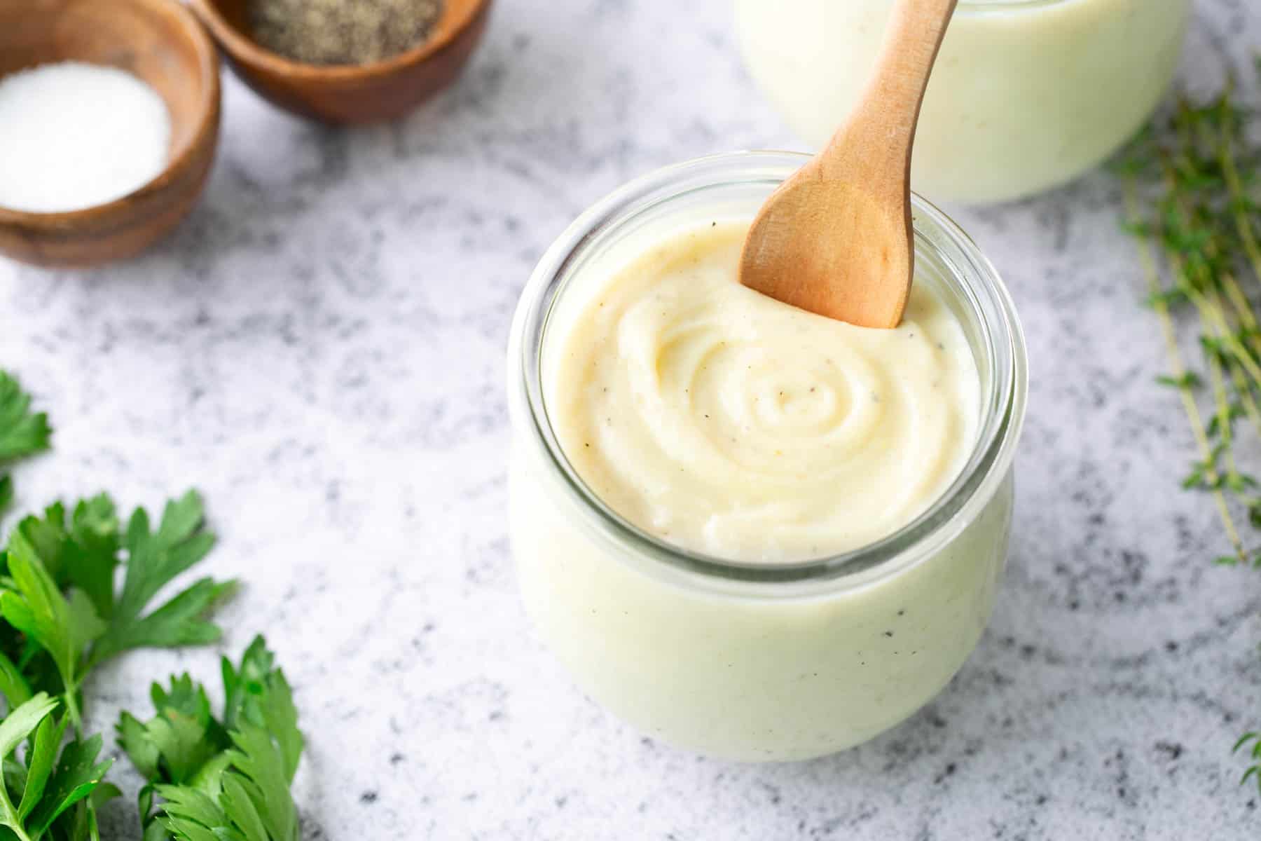 A glass jar filled with creamy white sauce, stirred with a wooden spoon, sits on a light countertop surrounded by fresh herbs and small bowls of salt and pepper.