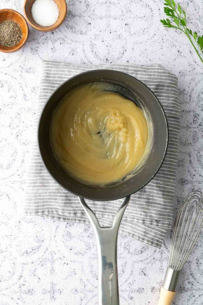 A saucepan containing a light-colored roux sits on a striped cloth, with a whisk nearby and small bowls of salt and pepper in the background.