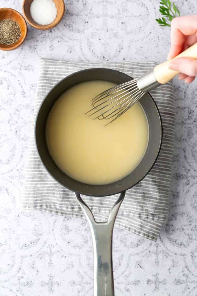 A hand whisking a light-colored sauce in a saucepan on a gray striped cloth, with small bowls of salt and pepper nearby.