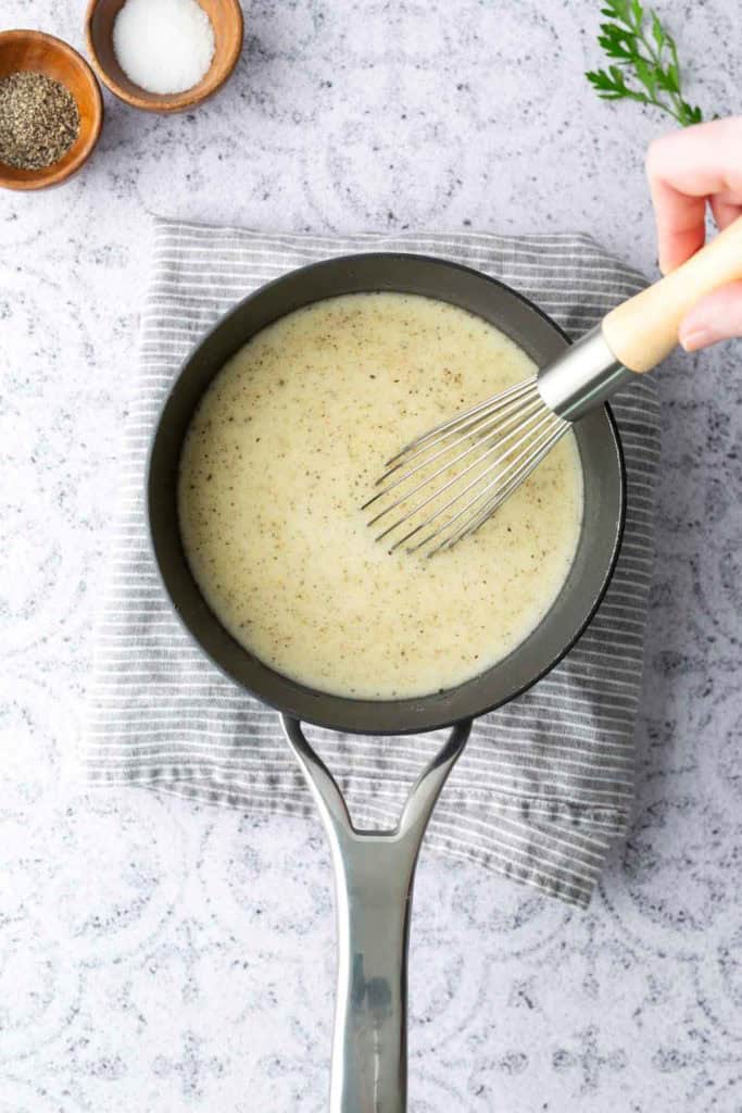 A hand whisks a creamy white sauce in a saucepan placed on a striped cloth, with bowls of salt and pepper and a sprig of parsley nearby.