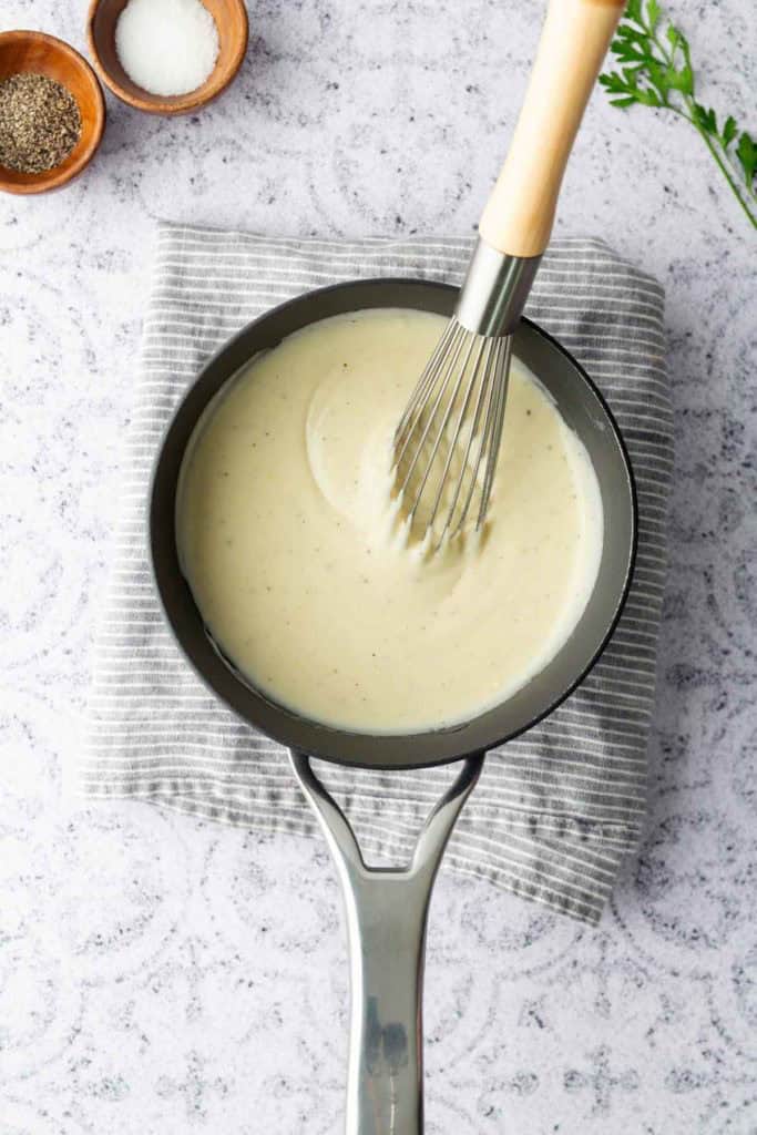 A saucepan filled with creamy white sauce being whisked, resting on a striped cloth with bowls of salt and pepper nearby.