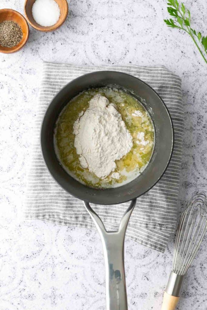 A saucepan containing melted butter and flour sits on a striped cloth, beside a whisk, salt, and pepper on a light countertop.