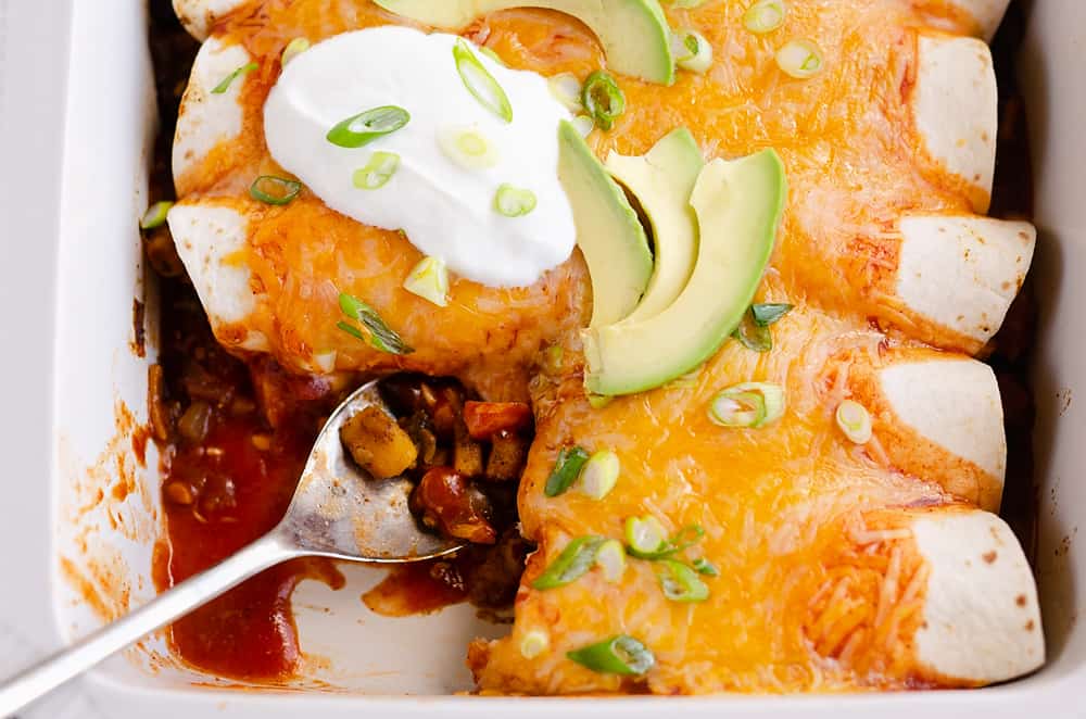 A baking dish with cheesy enchiladas topped with sour cream, avocado slices, and green onions. One enchilada is partially served with a spoon.