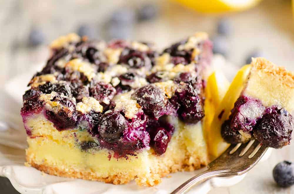 A close-up of a slice of blueberry dessert bar with a crumbly crust, creamy filling, and baked blueberries on top, with a fork holding a piece.