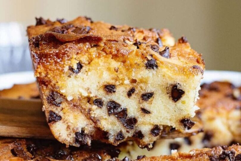 A close-up of a thick slice of cake with chocolate chips throughout, being lifted from the whole cake with a spatula.