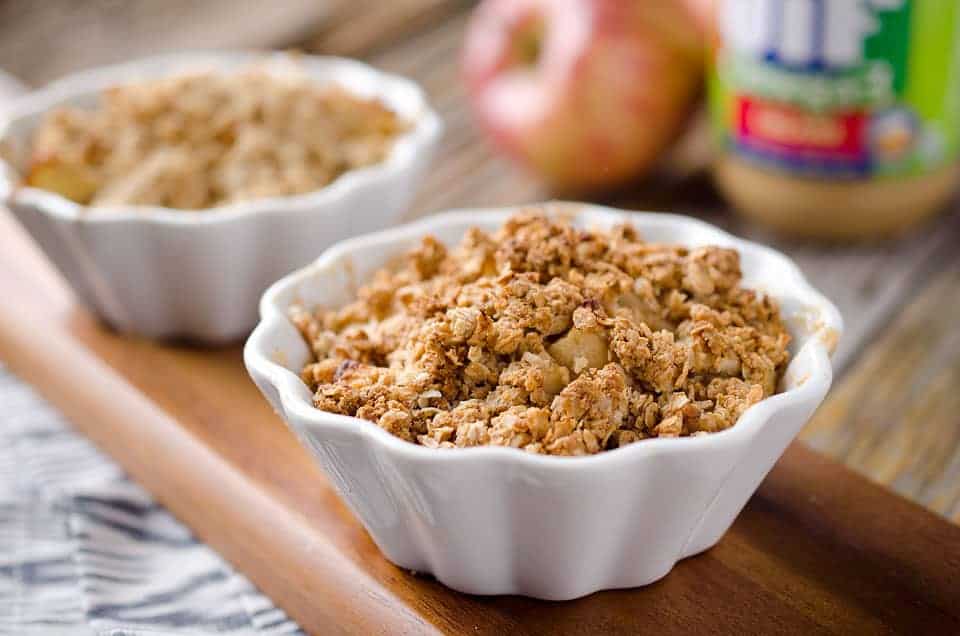Two white ramekins filled with baked apple crisp are placed on a wooden board, with an apple and a jar of peanut butter in the blurred background.