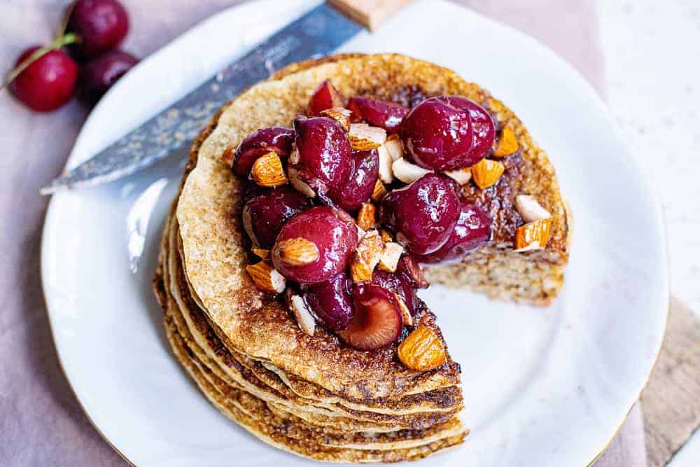 A stack of pancakes topped with fresh cherries and chopped almonds, with a slice cut out on a white plate next to a knife and some whole cherries.