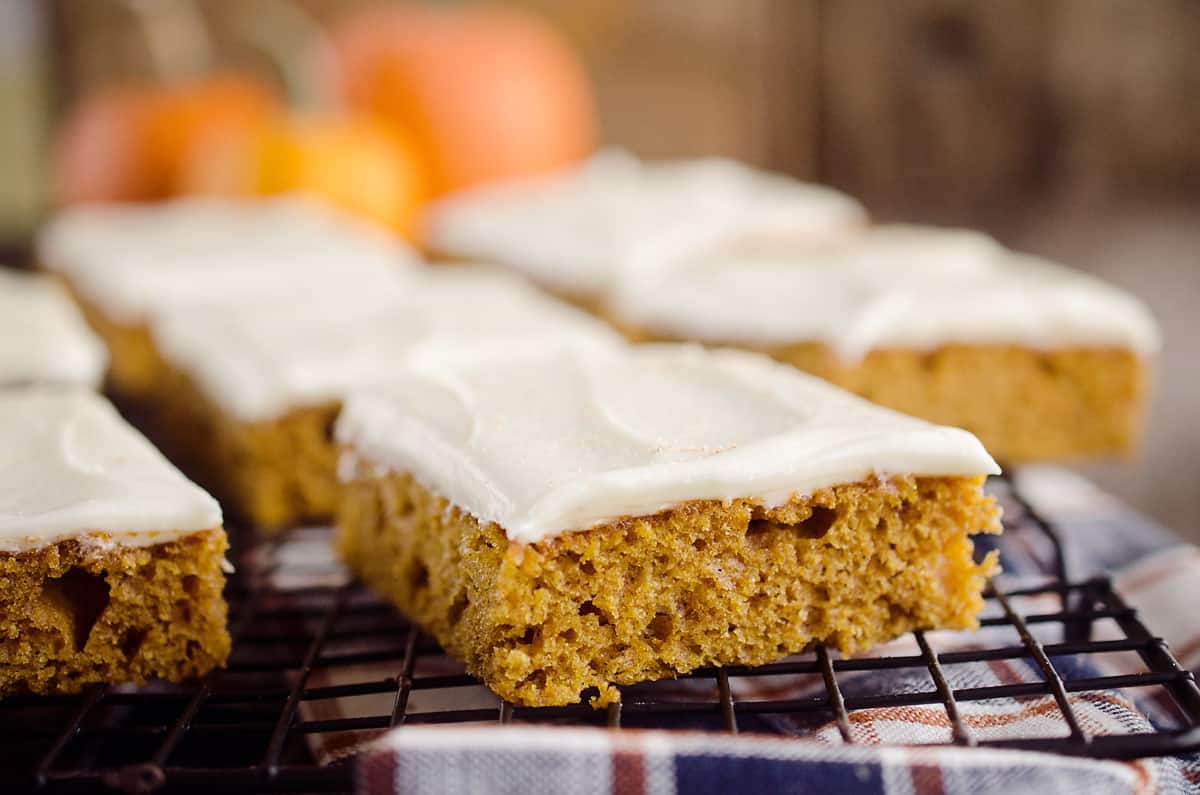Pumpkin bars with cream cheese frosting are arranged on a cooling rack, with a blurred background featuring orange shapes.