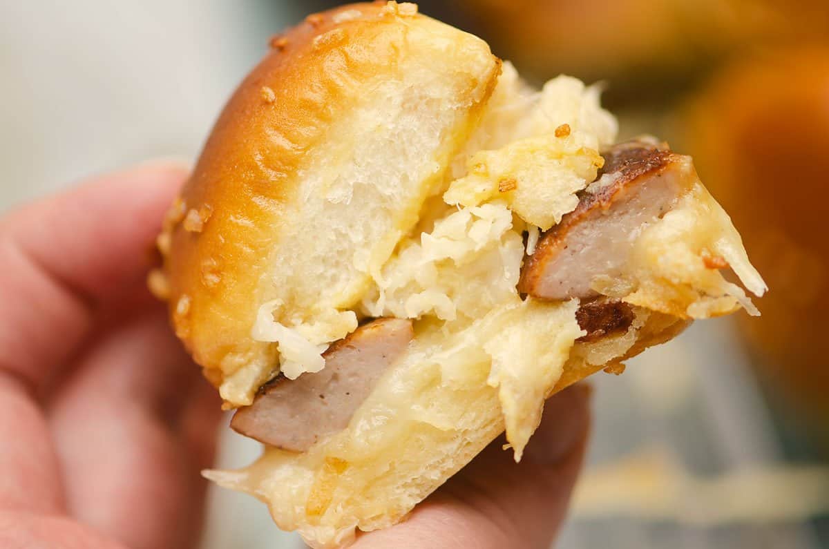 A close-up of a hand holding a sandwich with sliced sausage, sauerkraut, and melted cheese in a pretzel bun.