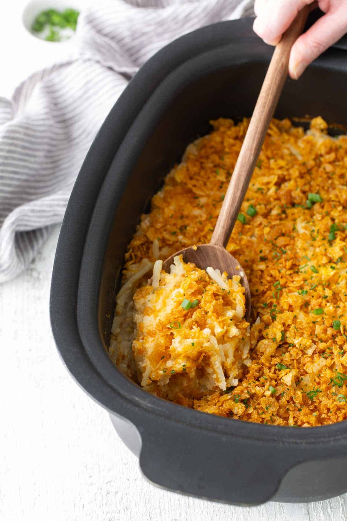 A hand uses a wooden spoon to serve a portion of hash brown casserole topped with a crispy crumb coating from a black baking dish.
