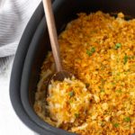 A black baking dish filled with hash brown casserole topped with a golden crumb crust, with a wooden spoon scooping out a portion.