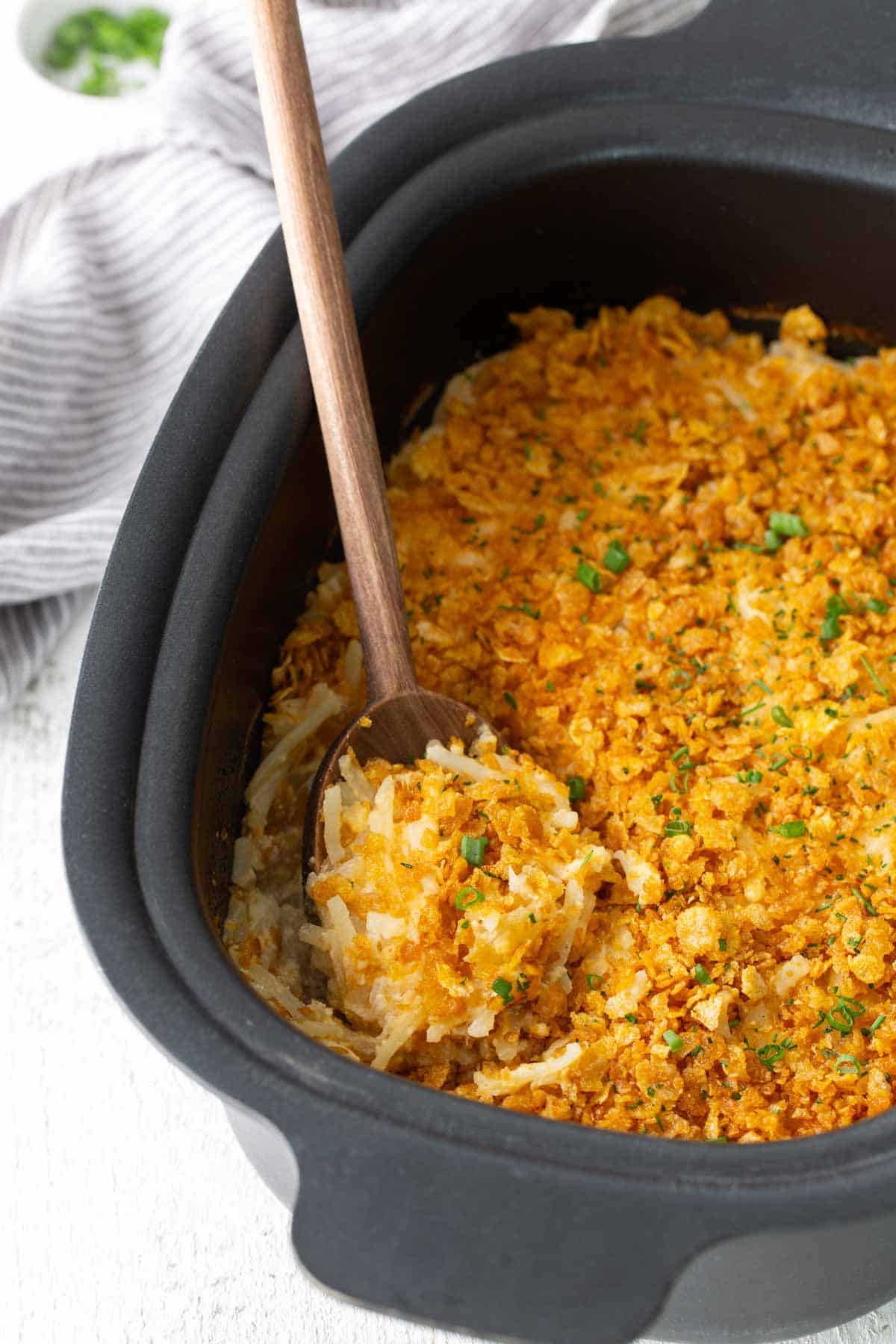 A black baking dish filled with hash brown casserole topped with a golden crumb crust, with a wooden spoon scooping out a portion.