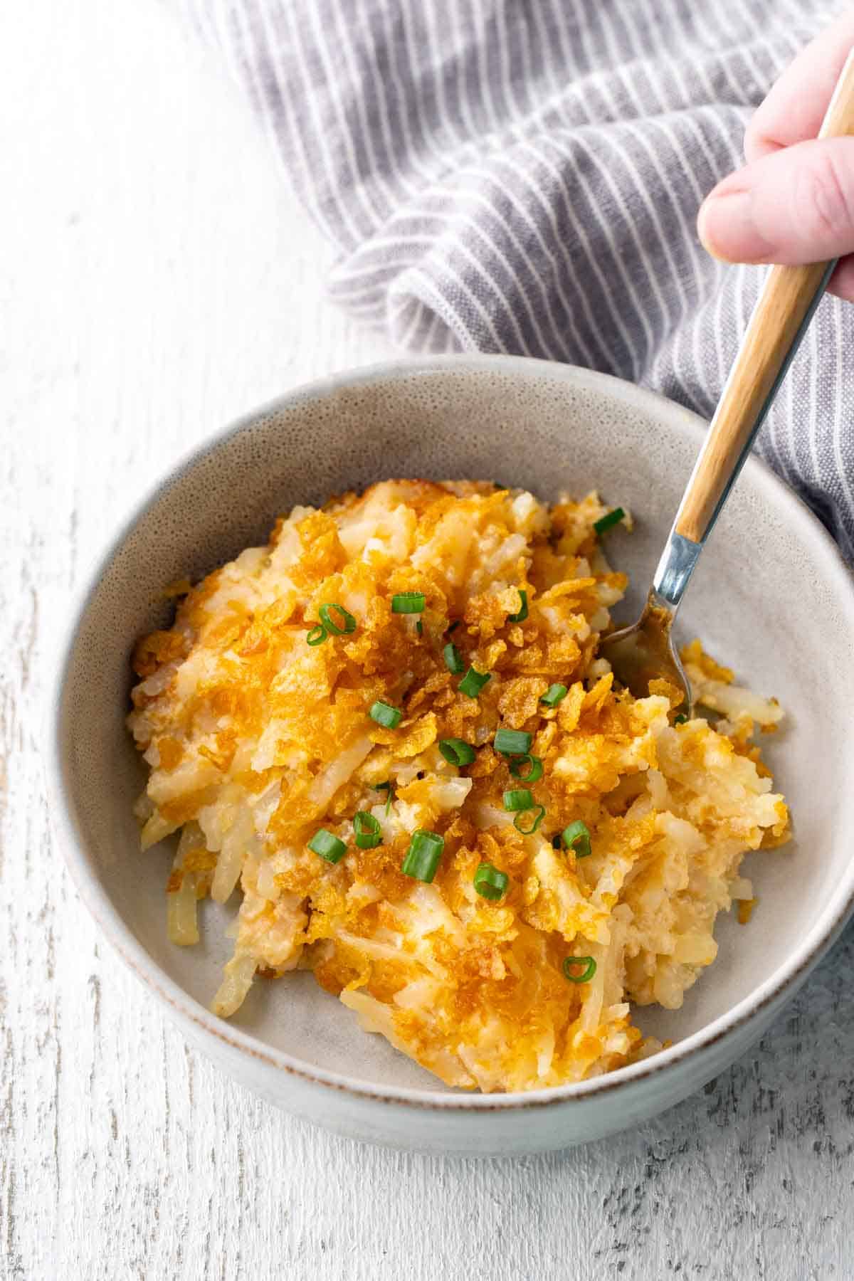 A bowl of cheesy hash brown casserole topped with crushed cornflakes and chopped green onions, with a fork and a striped cloth in the background.