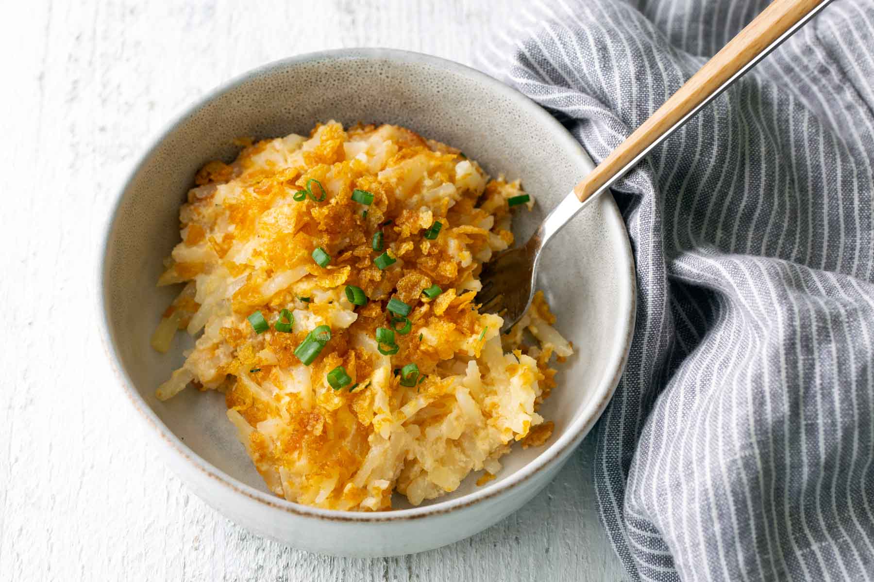 A bowl of baked hash brown casserole topped with chopped chives, with a fork and a gray striped cloth napkin beside it.