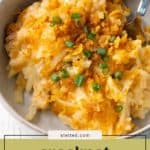 A bowl of crockpot cheesy potatoes topped with green onions and crispy crumbs, with a fork on the side.