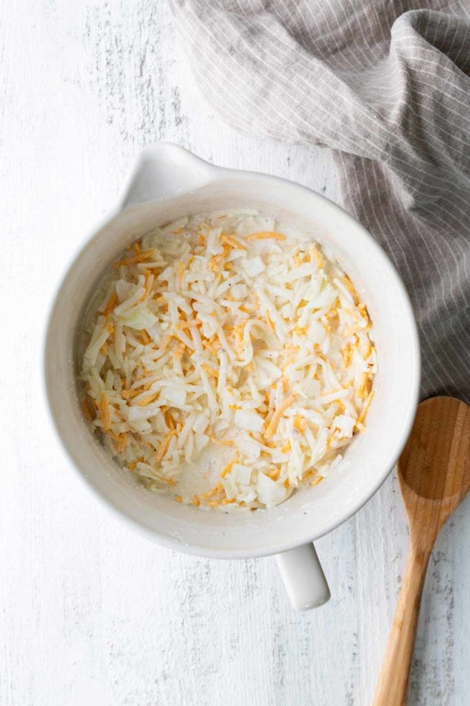 White mixing bowl with shredded potatoes and cheese mixture on a white surface, next to a wooden spoon and a gray striped cloth.