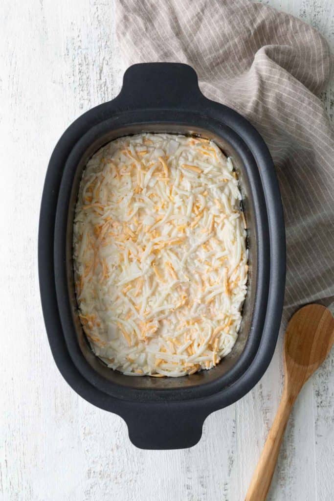 A black baking dish filled with a casserole topped with shredded cheese sits on a white surface next to a wooden spoon and a beige striped cloth.