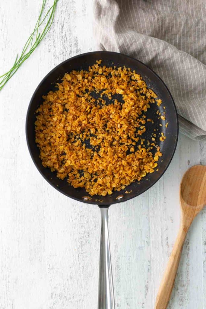 A black frying pan with cooked, seasoned cauliflower rice sits on a white surface next to a wooden spoon, a gray striped cloth, and a sprig of fresh herbs.
