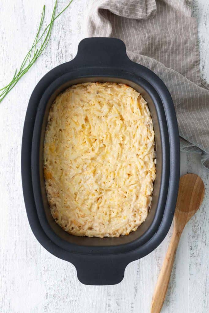 A baked casserole with a cheesy, golden top in a black dish, placed on a white surface next to a wooden spoon and a striped towel.