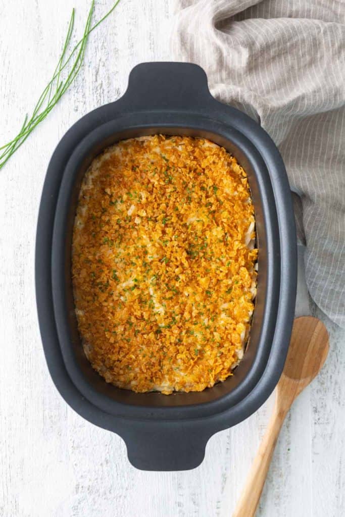 A black casserole dish filled with baked casserole topped with golden, crispy crumbs sits on a white surface next to a wooden spoon and a striped towel.