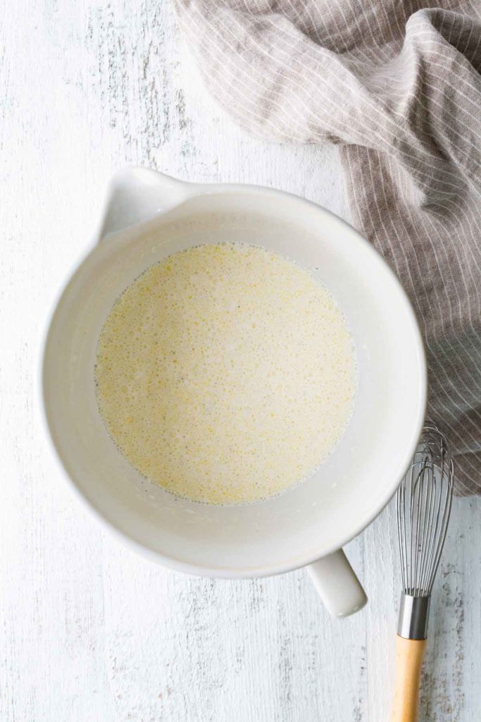 A mixing bowl with a pale, speckled liquid mixture inside, next to a metal whisk and a striped kitchen towel on a white surface.